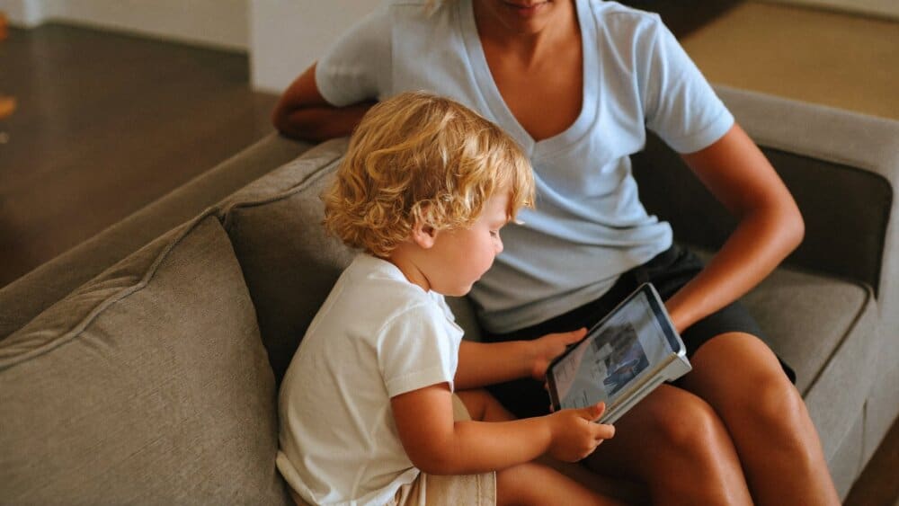 A parent supervising a young child using a tablet together on the couch