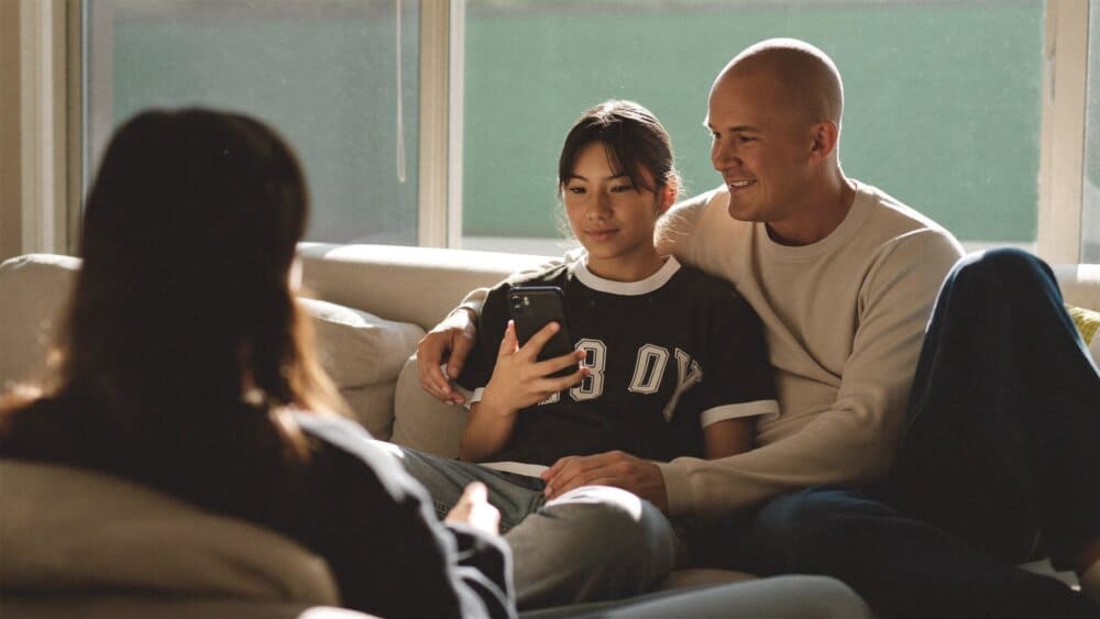 A parent and teenage child sitting on a living room couch talking calmly while the teen holds a phone