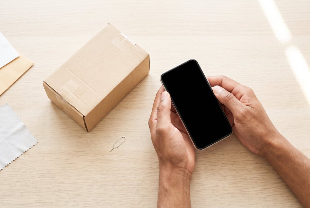Hands holding a smartphone with a blank screen next to a shipping box, preparing to sell a phone
